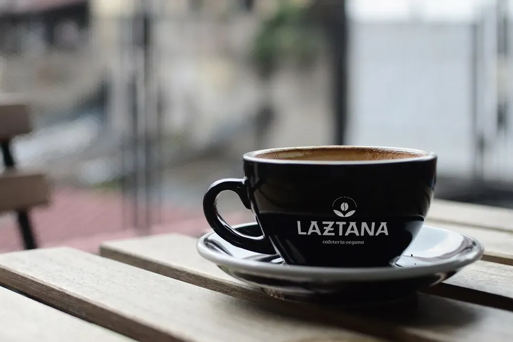 Taza negra de café con el logotipo de una cafetería vegana en Bilbao en blanco.